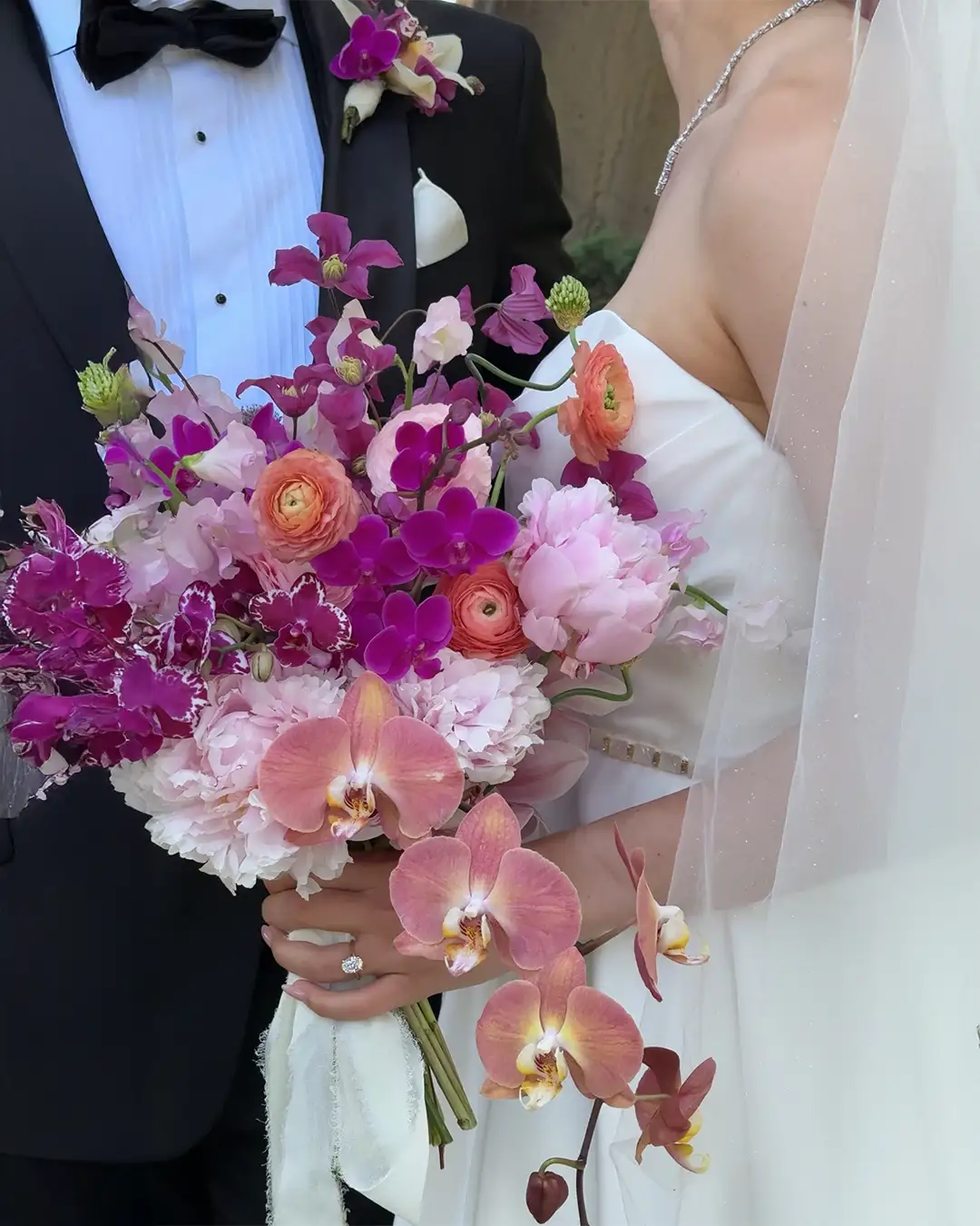 Gelin ve damadın yakın çekim görüntüsünde, mor orkideler, leylak rengi clematis, şeftali ranunculus ve pembe şakayıkların bir araya geldiği, beyaz kurdeleyle bağlanmış gösterişli ve modern bir mor gelin çiçeği buketi gelinin elinde parıldayan pırlanta yüzükle dikkat çekiyor.