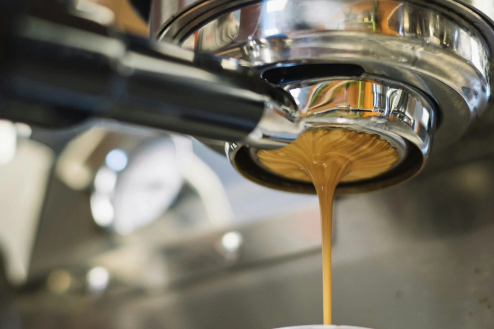 Bir espresso kahve makinesinin parlak krom portafiltresinden, kalın ve kremsi altın kahverengi espresso kahvesi bir fincana doğru akarken çekilen bu detaylı yakın çekimde, kahve akışının görsel yoğunluğu ve makine parçalarının yansımaları belirgindir.