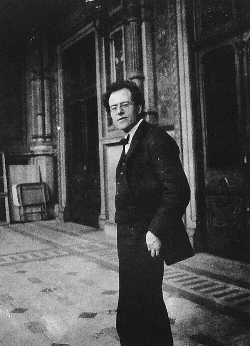 1903 tarihli siyah beyaz fotoğrafta, koyu renk takım elbisesi ve gözlükleriyle ciddi bir ifadeyle bize doğru bakan Gustav Mahler, Viyana Operası'nın ihtişamlı iç mekanında duruyor, arkasında süslü mimari detaylar ve solunda bir sandalye seçiliyor.