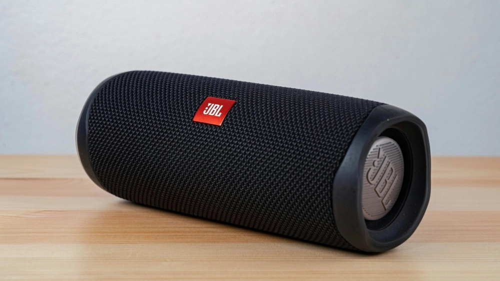 Ahşap bir yüzeyde duran, koyu renkli, dokulu bir ızgaraya ve belirgin kırmızı JBL logosuna sahip silindir şeklindeki taşınabilir bir kablosuz Bluetooth hoparlör, makalenin ana konusunu oluşturan modern ses teknolojisini vurguluyor.