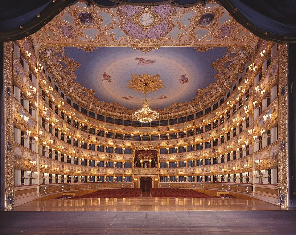 Venedik'teki La Fenice Opera Binası'nın görkemli iç mekanı, altın varaklı ve detaylı oymalı çok katlı locaları, sahneyi çevreleyen ışıklı yapısıyla, melek figürleriyle bezenmiş mavi tavan freski ve ortasındaki büyük kristal avizeyle tarihi ihtişamını gözler önüne seriyor.