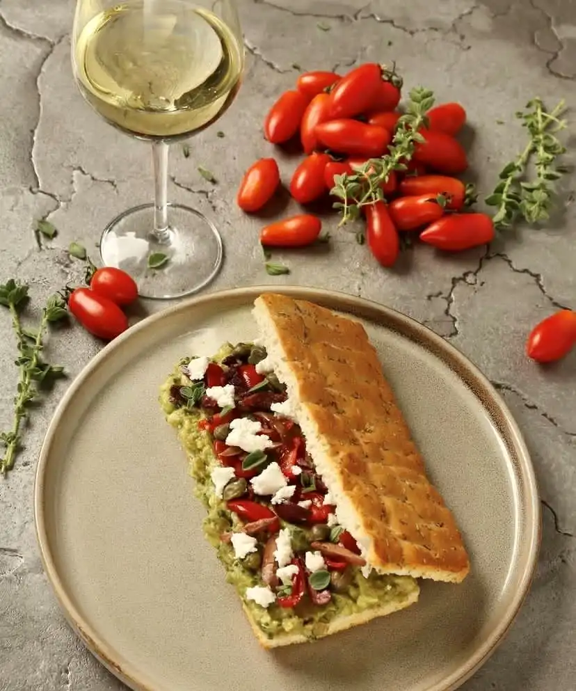 Gri bir zeminin üzerinde duran tabakta, yeşil avokado veya benzeri bir ezme, ufalanmış beyaz peynir, kırmızı biber ve kapari ile zenginleştirilmiş, üzerine taze kekik serpilmiş modern bir ekmek arası sunum yer alıyor; yanında bir kadeh beyaz şarap ve bolca taze çeri domates bulunan bu kare, Selanik'in çeşitli ve çağdaş gastronomik lezzet duraklarından birine ait, iştah açıcı bir öğünü gözler önüne seriyor.