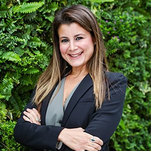 Orta uzunlukta kahverengi saçlara sahip, açık yeşil bir bluz ve koyu renk bir blazer ceket giyen, kolları kendinden emin bir şekilde kavuşturulmuş, gür yeşil bir bitki duvarının önünde içtenlikle gülümseyen bir kadın portresi.