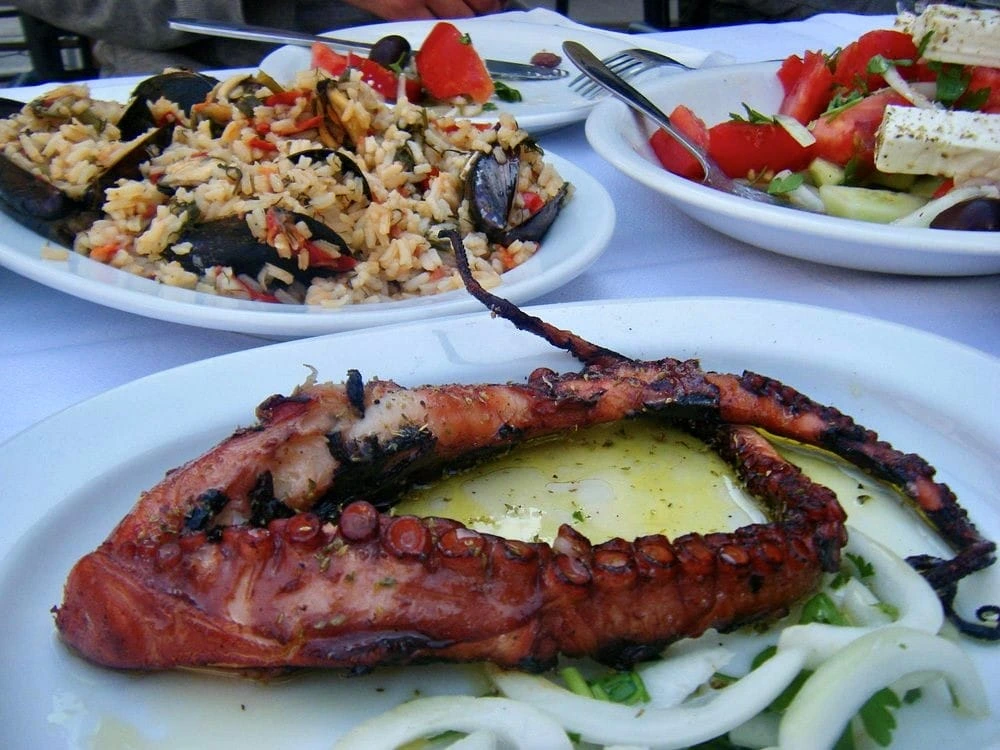 Selanik'in en iyi restoranları listesinde yer alan taze deniz ürünleri gastronomisine özgü bir ziyafet masası; ortasında zeytinyağıyla parlaklaşan ızgara ahtapot, midyelerle zenginleştirilmiş pilav ve küp beyaz peynirli Akdeniz salatasıyla göz alıcı bir şekilde sunuluyor.