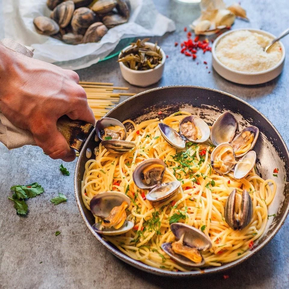 Bir elin tavayı tuttuğu karede, üzerinde taze istiridyeler, doğranmış yeşillikler ve kırmızı biber parçacıklarıyla süslenmiş lezzetli spaghetti vongole tabağı Selanik'in en iyi restoranlarında bulunabilecek zengin deniz ürünleri seçeneklerini temsil ediyor, arka planda çiğ istiridyeler, turşular ve rendelenmiş peynir görülüyor.