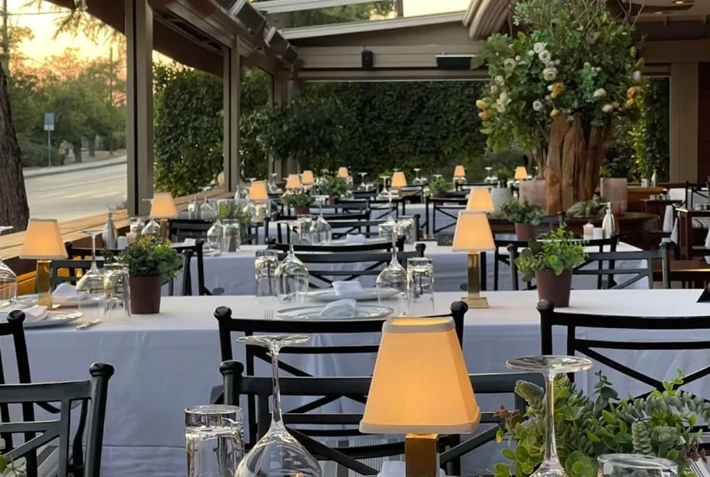 Beyaz örtülü masalar, zarif masa lambaları ve yeşil bitki süslemeleriyle davetkar bir atmosfere sahip, Selanik'in en iyi restoranlarından Mavri Thalassa'nın açık hava yemek alanı akşam için hazır.