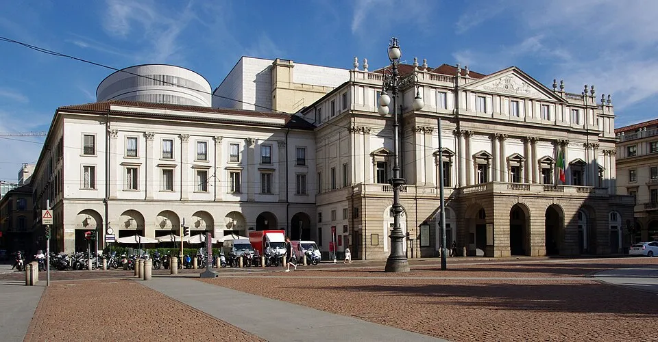 Milano'daki ünlü La Scala opera binası, geniş bir taş meydanın kenarında yer alırken, bej ve beyaz tonlarındaki klasik sütunlu cephesiyle dikkat çekiyor ve sol arka tarafında modern bir ek yapı göze çarpıyor.