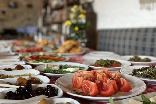 Çanakkale Seyahat Rehberi'nde bahsettiğimiz gibi, yöresel ürünlerle hazırlanmış zengin bir kahvaltı sofrası: bolca dilimlenmiş domates, zeytin, peynir ve otlu mezelerin bulunduğu beyaz tabaklar kırmızı-beyaz ekose örtülü masada iştah açıcı bir şekilde sergilenirken, arka planda sıcak ekmekler ve sarı çiçekler seçiliyor.