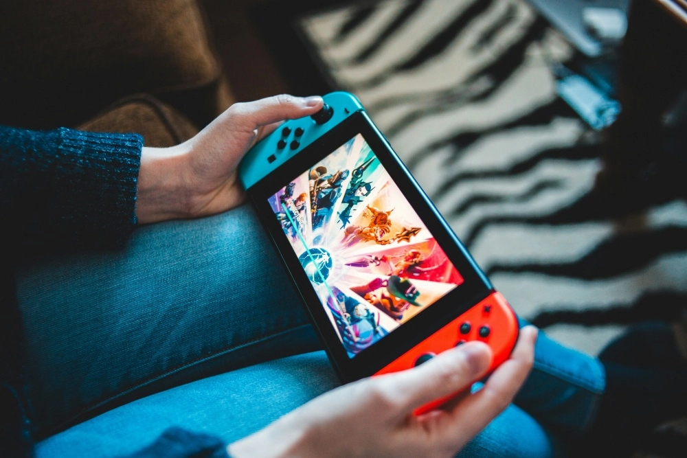 Mavi kot pantolonlu birinin ellerinde, çok sayıda aksiyon karakterinin canlı renklerle gösterildiği bir oyunun açık olduğu ekranıyla neon mavi ve neon kırmızı Joy-Con'lara sahip Nintendo Switch el oyun konsolu yakın çekimde görülüyor.