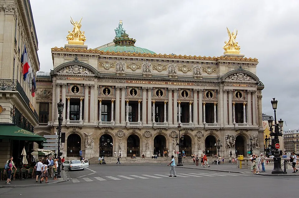 Paris'teki görkemli Opéra Garnier'nin altın heykellerle süslü, yeşil kubbeli ve detaylı klasik mimariye sahip ön cephesi, bulutlu bir günde, önünde yayaların ve bir kafenin bulunduğu kalabalık bir caddeden görünmektedir.
