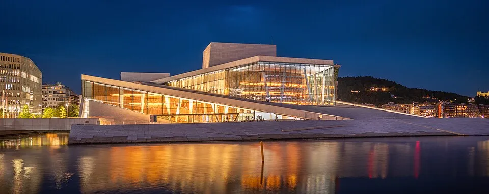 Karanlık gökyüzü altında, Oslo'nun sularında yansıyan ışıklarıyla Oslo Opera Binası, beyaz mermerden eğimli çatıları ve içinden yayılan sıcak bir parıltıyla aydınlanan cam cephesiyle modern bir mimari harikasıdır.