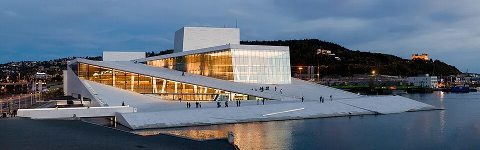 Alacakaranlıkta parlayan modern mimarinin bir örneği olan Oslo Opera Binası, denize doğru eğimli beyaz mermer çatısı üzerinde yürüyen veya dinlenen birçok insanla canlı bir kamusal alan sunarken, arkasında şehir ışıkları ve uzak bir tepedeki kale silüeti belirginleşiyor.