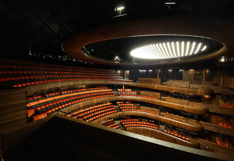 Oslo Opera Binası'nın içinden çekilmiş bu geniş açılı görselde, boş, kıvrımlı sıralar halinde dizili, alttan turuncu ışıkla aydınlatılmış modern koltuklar ve ahşap detaylarla süslü balkonlar görülüyor; tavanı aydınlatan dairesel, çizgisel lambalar mekanın çağdaş tasarımını vurguluyor.