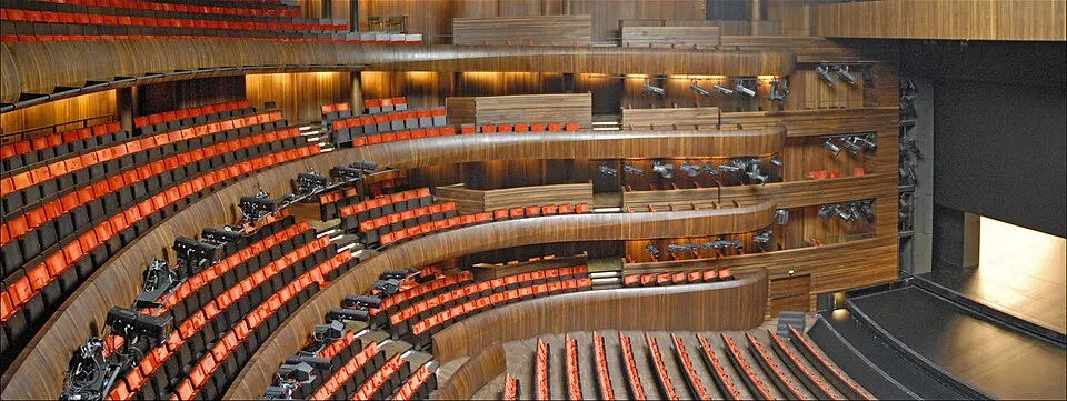 Oslo Opera Binası'nın içini gösteren bu geniş açılı görüntüde, sahneye doğru kavisli olarak uzanan turuncu ve koyu renkli koltuk sıraları, çok katlı ahşap balkonlar ve sahne ışıkları, modern ve davetkar bir opera salonu atmosferi sunuyor.
