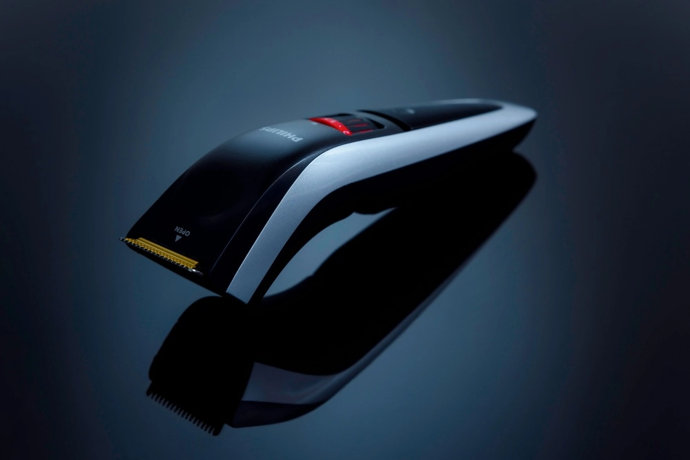 Siyah ve gümüş renk tonlarına sahip, modern tasarımlı, şarjlı bir Philips tıraş makinesi, parlak koyu zeminde net yansımasıyla belirginleşirken, kırmızı ışıklı göstergesi ve altın renkli kesici bıçakları saç ve sakal için ayarlanabilir başlık özelliğini öne çıkarıyor.