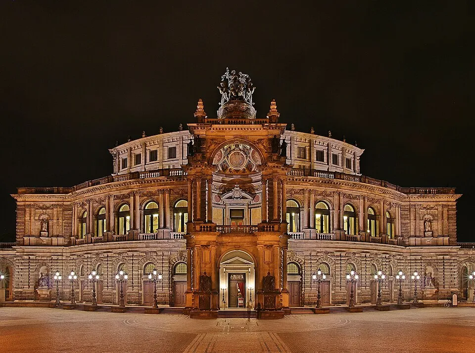 Gece karanlığında, Dresden'deki görkemli Semperoper binası, çatısındaki heykelleri, kemerli pencerelerinden sızan sıcak ışıkları ve önündeki geniş meydanı aydınlatan süslü sokak lambalarıyla pırıl pırıl parlayarak ziyaretçilerini ağırlayan bir sanat ve kültür yapısıdır.
