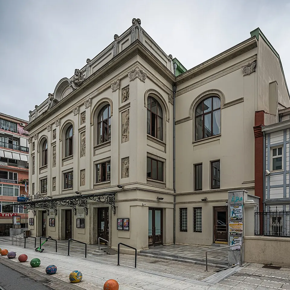 Gri bulutlu bir günde, kemerli pencereleri, kabartma süslemeleri ve ferforje kanopisiyle dikkat çeken Süreyya Operası'nın dış cephesi, önündeki renkli top şeklindeki yol sınır taşları ve bir scooter ile birlikte modern şehir yaşamına tanıklık eden tarihi bir yapı olarak görünüyor.