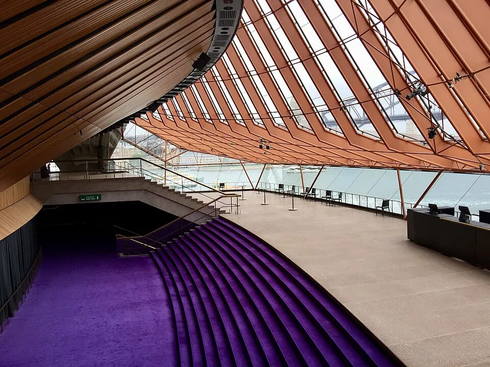 Sydney Opera Evi'nin içindeki geniş salonda, denize bakan eğimli pencerelerden Sidney Liman Köprüsü görünürken, solda mor kadife basamaklı kıvrımlı bir merdiven, yukarıda ise ahşap panelli zarif tavan dikkat çekiyor.