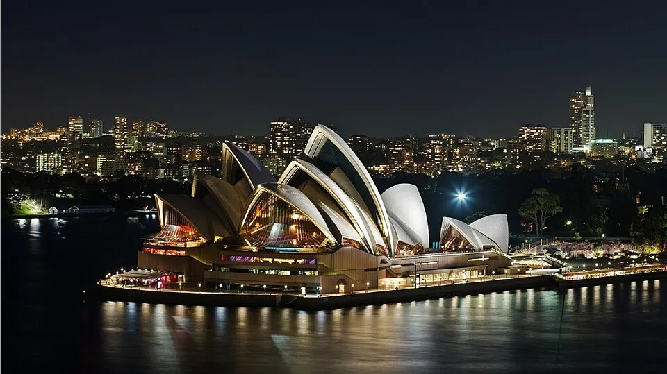 Sydney Opera Binası, karanlık gece gökyüzünün altında, kendine özgü yelken benzeri çatıları parlak sarı ve beyaz ışıklarla aydınlatılmış halde sudan yükseliyor, arka planda ise şehir silüetinin pırıltılı ışıkları uzanarak bu ikonik yapının büyülü gece atmosferini tamamlıyor.