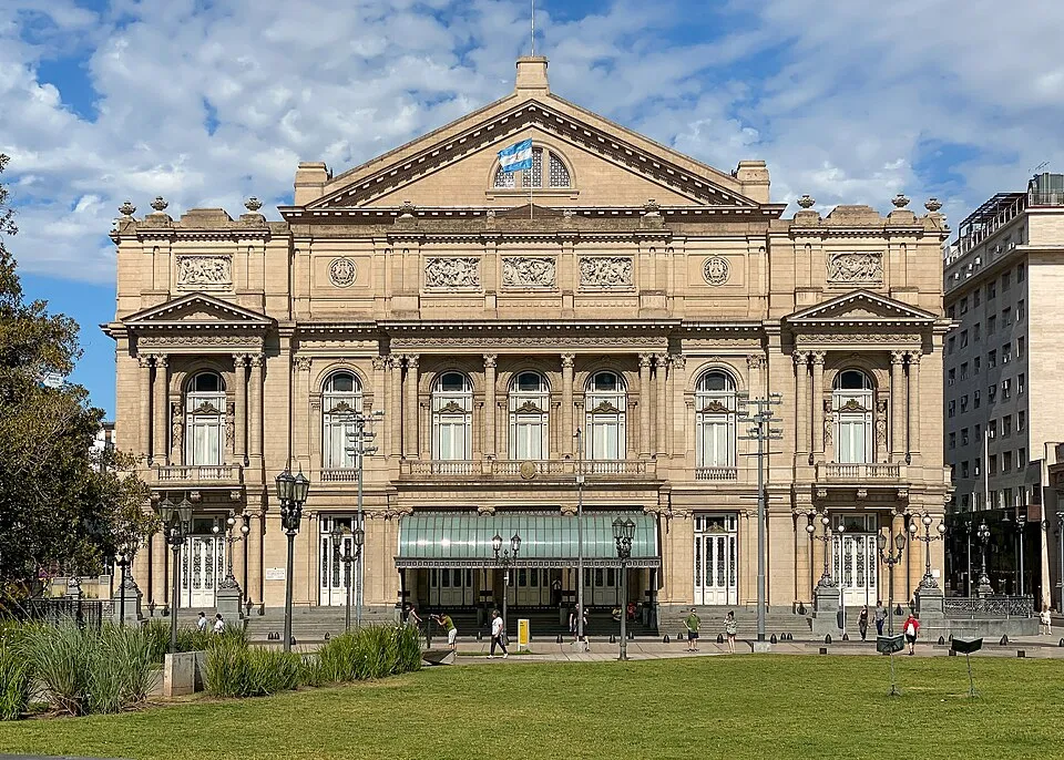 Büyük, gösterişli ve klasik mimariye sahip, tepesinde Arjantin bayrağı dalgalanan, bej renkli Teatro Colón opera binası, önünde yeşil bir çim alan ve dolaşan insanlarla birlikte Buenos Aires'in merkezinde güneşli bir günde görünüyor.