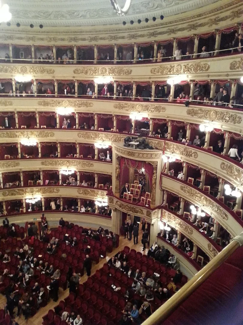 Milan'daki ünlü Teatro alla Scala'nın loggione bölümünden görülen bu etkileyici iç mekanda, kırmızı kadife koltuklarda oturan ve gösteri için hazır bekleyen kalabalık seyirciler, altın varak süslemeli tarihi salonun ihtişamını vurguluyor.