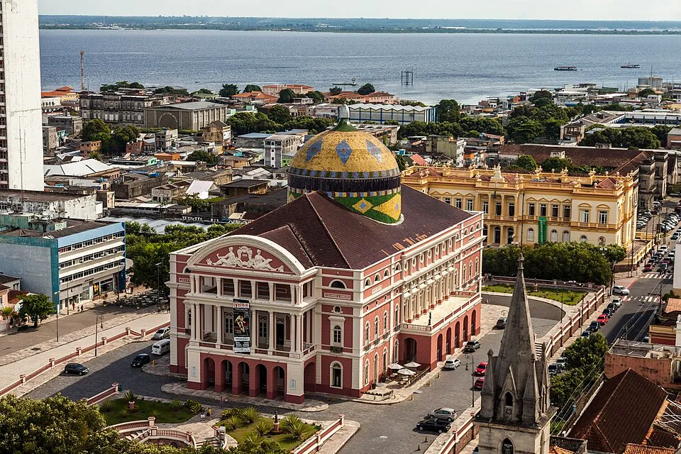 Manaus'un ortasında yer alan, kırmızımsı pembe cephesi, beyaz sütunları ve Brezilya bayrağının canlı renklerini yansıtan mavi, sarı, yeşil desenli kubbesiyle dikkat çeken görkemli Teatro Amazonas Opera Binası, çevresindeki yoğun şehir yapıları ve uzakta parlayan geniş nehirle birlikte kuşbakışı gösterilmektedir.