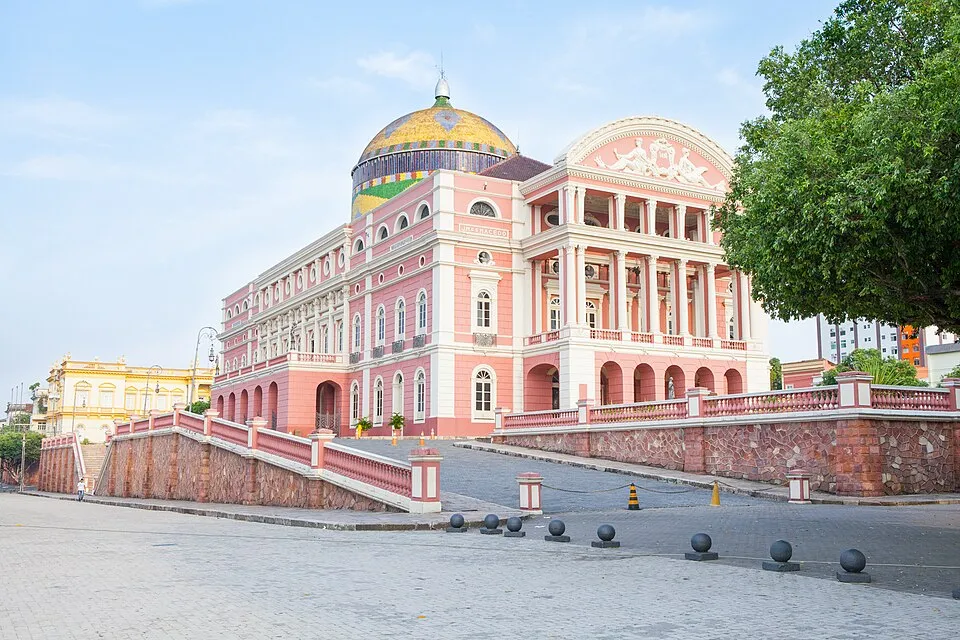 Açık pembe ve beyaz neoklasik tarzda, görkemli sütunları, kemerleri ve balkonları olan, altın ve mozaik desenli büyük bir kubbeyle taçlanmış Teatro Amazonas opera binası, önündeki geniş meydan ve sağındaki yeşil ağaçla birlikte gün ışığında uzaktan görünüyor.