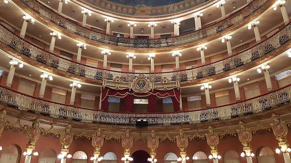Teatro Amazonas'ın içinden yukarı doğru bakışla çekilmiş bu görüntüde, altın yaldızlı detaylarla süslü kavisli balkonlar ve aydınlatma armatürleri, sahneye bakan ana locanın ihtişamını vurgulayan kırmızı perdeler ve zarif oymalarla bir opera binasının zengin mimarisini sergiliyor.