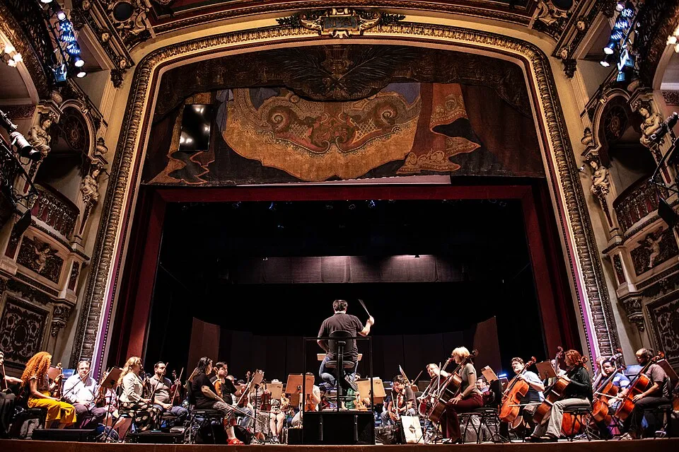 Teatro Amazonas'ın görkemli opera sahnesinde, orkestra şefinin yönetiminde birçok müzisyenin yaylı enstrümanlarını çalarak prova yaptığı veya performans sergilediği, sahne arkasında ve yanlarda altın varak işlemeli süslemelerin ve antika detayların göz kamaştırdığı geniş açılı bir görünüm.