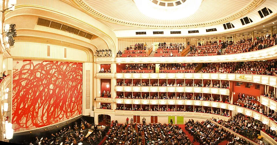 Viyana Devlet Operası'nın görkemli salonunda, sahne önünde canlı orkestra ve kırmızı soyut fırça darbelerinden oluşan büyük bir sanat eseri sahne perdesi olarak dururken, altın rengi detaylarla süslü balkonlar ve localar tüm katlarda şık giyimli seyircilerle dolup taşıyor.
