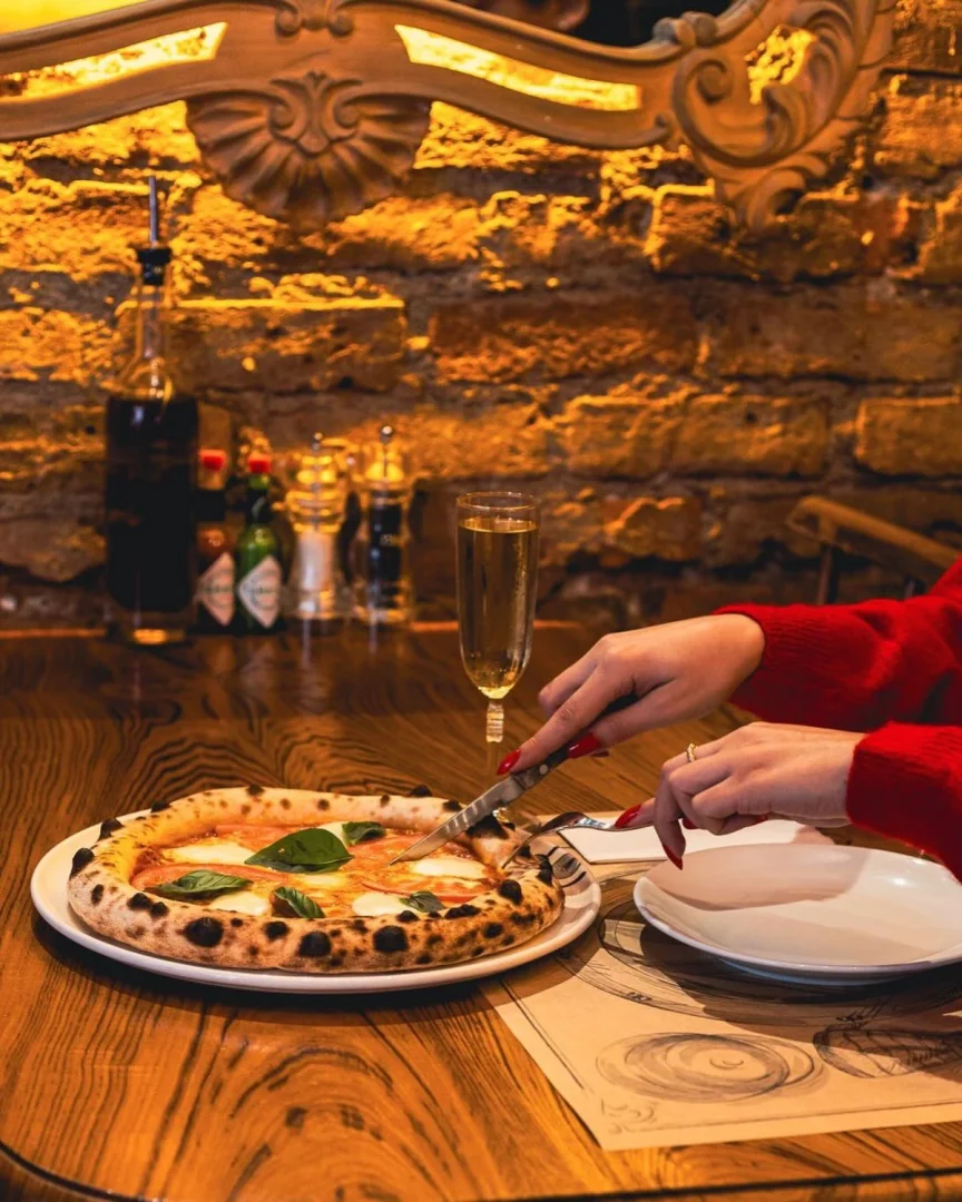 Kırmızı kazaklı, tırnakları ojeli bir kişinin ahşap masada, üzerinde taze fesleğen ve mozzarella peyniri olan, kenarları hafifçe yanmış Napoliten tarzı bir pizzayı bıçakla keserken yakından görüldüğü bu karede, arka plandaki loş ışıklı tuğla duvar ve zarif ayna detayı sıcak bir restoran atmosferi sunuyor.