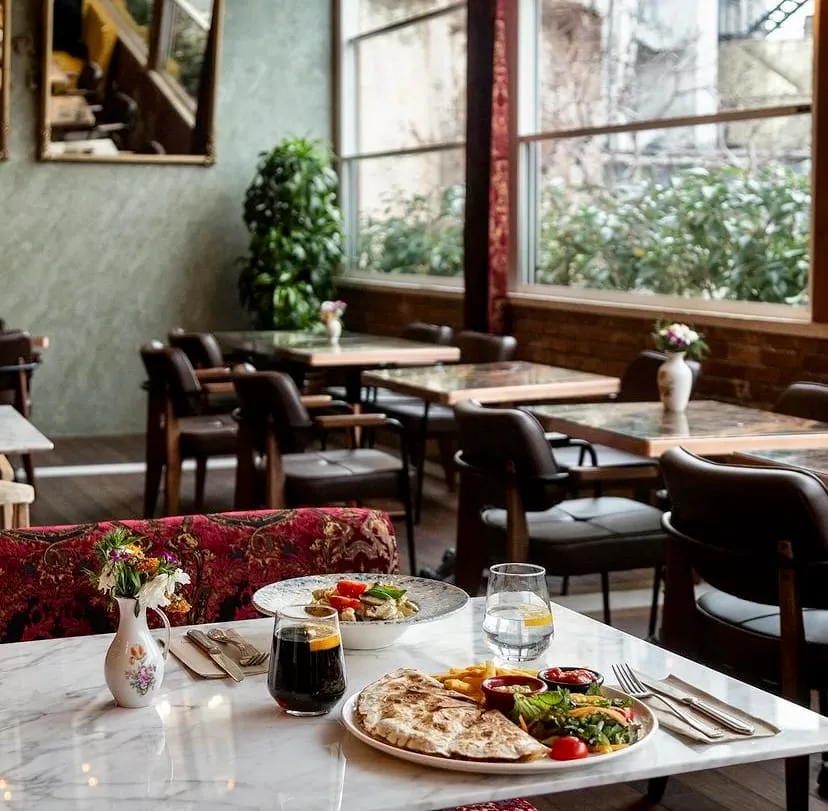 Beyaz mermer bir masanın üzerinde, patates kızartması, salata ve pide gibi çeşitlerin bulunduğu zengin bir ana yemek tabağı ile yanında domatesli makarnalı bir ara yemek, iki içecek ve taze çiçekler dikkat çekerken, loş ışıklı arka planda boş masalar ve büyük pencerelerden dışarıdaki yeşil bitkilerle aydınlanan ferah bir restoran ortamı.
