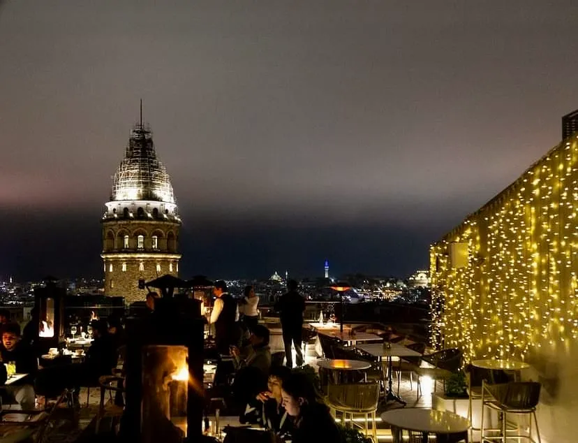 Galata Kulesi'nin ışıklı silüetiyle süslenen gece manzarasında, iskeleli tepesiyle yükselen kuleye karşı konumlanmış, sarı peri ışıklarıyla bezeli bir çatı katı barında oturan ve ayakta duran insanlar, şık masalarda içeceklerini yudumlayıp sohbet ederken, arkada İstanbul'un şehir ışıkları parlıyor.
