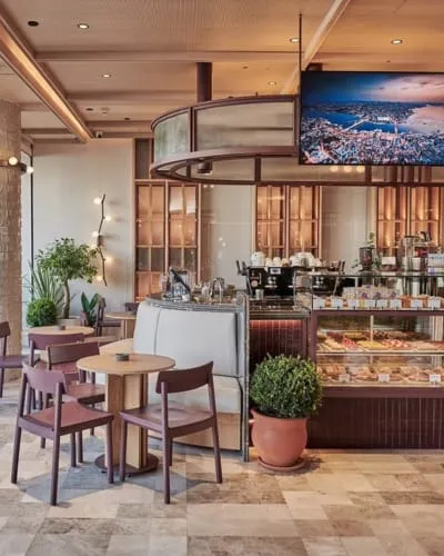 Ahşap tavan ve bitki detaylarıyla modern ve sıcak bir atmosfere sahip kafenin iç mekanında, ön planda kahverengi yuvarlak masalar ve koyu renk sandalyeler yer alırken, arka planda kahve makinelerinin bulunduğu bir bar tezgahı ve pastaların sergilendiği bir vitrin görünmektedir.
