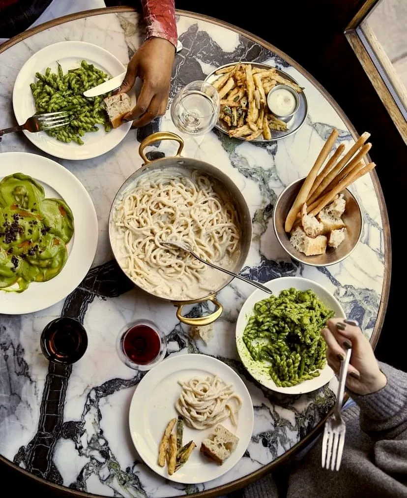 Mermer bir masanın üzerinde, ortada büyük bir tencerede kremalı spagetti, farklı tabaklarda yeşil pesto soslu makarnalar, yeşil mantılar, kızarmış patates ve yanında sosu, grissini ve ekmek dilimleri, iki kadeh kırmızı şarap ve bir bardak su içeren, birden fazla kişinin aynı anda yemek yediği zengin bir masa düzeni kuşbakışı görünüyor.