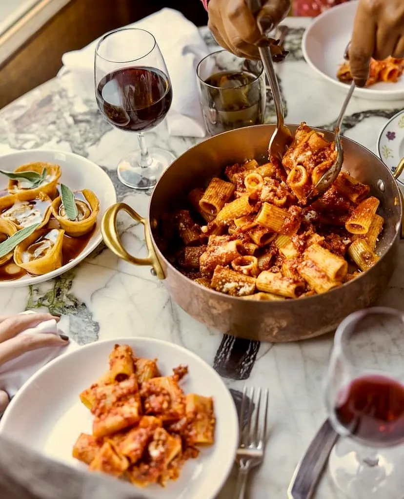 Ortasında bakır bir tencerede bolonez soslu rigatoni makarna bulunan, mermer bir masada, bir elin büyük servis kaşıklarıyla tencereden makarnayı beyaz bir tabağa aktardığı, sol üstte kırmızı şarap kadehi ve solda adaçayı yapraklı tartoletler ile dolu bir diğer tabağın görüldüğü, zengin bir İtalyan yemeği ziyafetini yansıtan samimi bir masa düzeni.