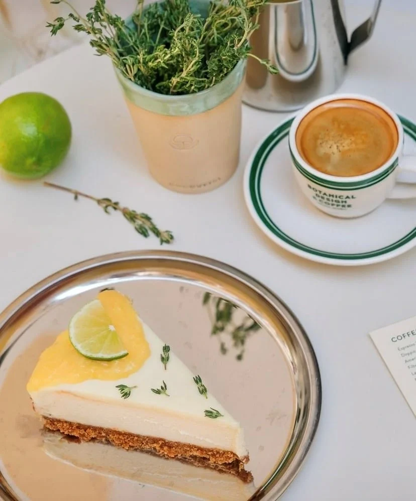 Bir dilim misket limonlu ve kekik süslemeli cheesecake, yanında BOTANICAL DESIGN COFFEE yazılı fincanda sıcak kahve, taze bitkilerle dolu bir saksı ve bütün bir misket limonu beyaz bir masanın üzerinde özenle duruyor.