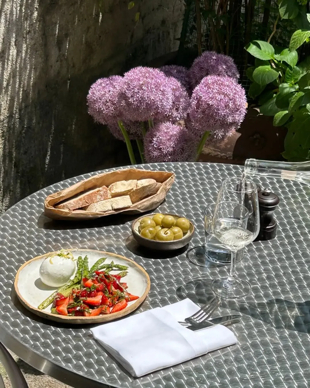 Güneşli havada, dış mekanda, dokulu metal bir masada, arka planda belirgin mor allium çiçeklerinin bulunduğu keyifli bir yemek düzenlemesi; taze burrata peyniri, kuşkonmaz ve çileklerin eşlik ettiği canlı bir salata, yanında ekmek, zeytin ve kadehte beyaz şarap görülüyor.