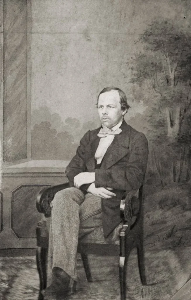 Dostoyevsky'nin hayatı ve eserleri hakkında bilgi veren yazıya eşlik eden bu fotoğraf, sandalyede oturan, arkasında hafif ağaçlı ve bulutlu bir fonun olduğu, ciddi bir ifadeyle bakan, papyonlu ve koyu renk takım elbiseli yazarın siyah beyaz bir portresidir.