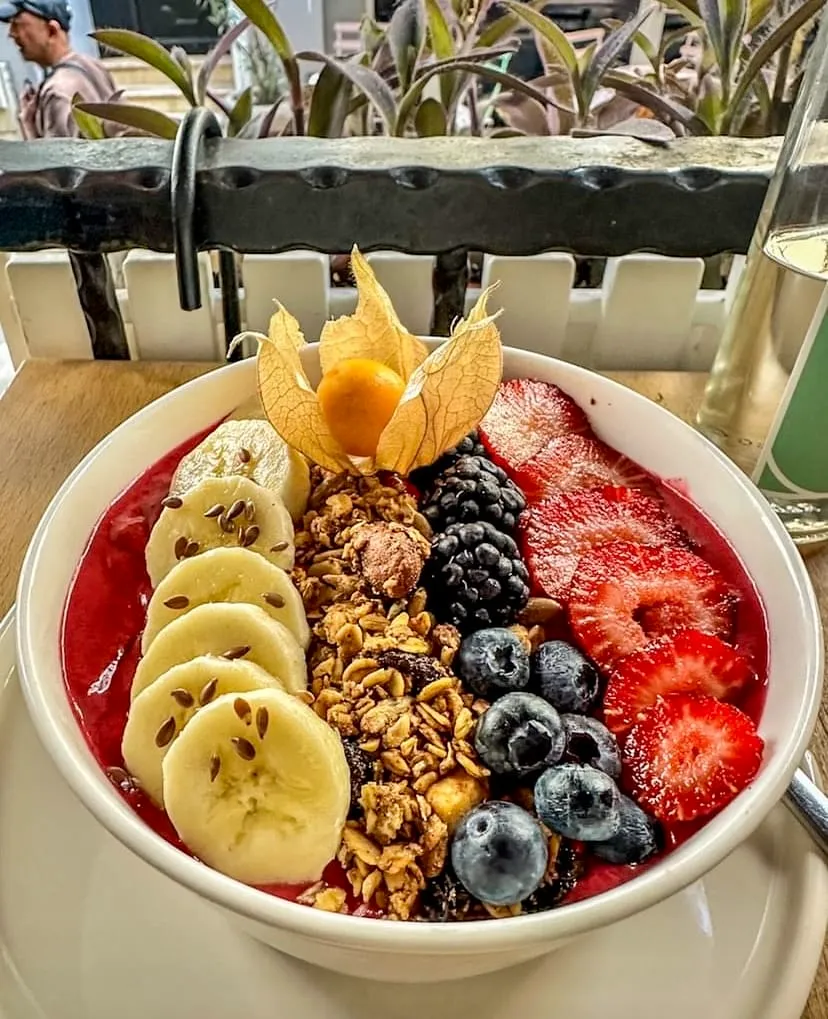 Beyaz bir kasede servis edilen, kırmızı renkli smoothie tabanının üzeri dilimlenmiş muz, keten tohumu, granola, böğürtlen, yaban mersini, çilek dilimleri ve ortasında açılmış bir altıntop ile bolca süslenmiş, arka planda bulanık yeşil bitkiler ve beyaz çit detayı görünen iştah açıcı bir kase.