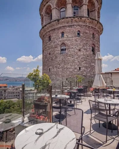 Güneşli bir çatı terası restoranı, sağ tarafta tarihi taş Galata Kulesi'nin belirgin bir şekilde yükseldiği, uzakta deniz ve şehir binalarının mavi gökyüzü altında net bir şekilde görüldüğü, birçok masa ve sandalyeye sahip bir manzara sunuyor.