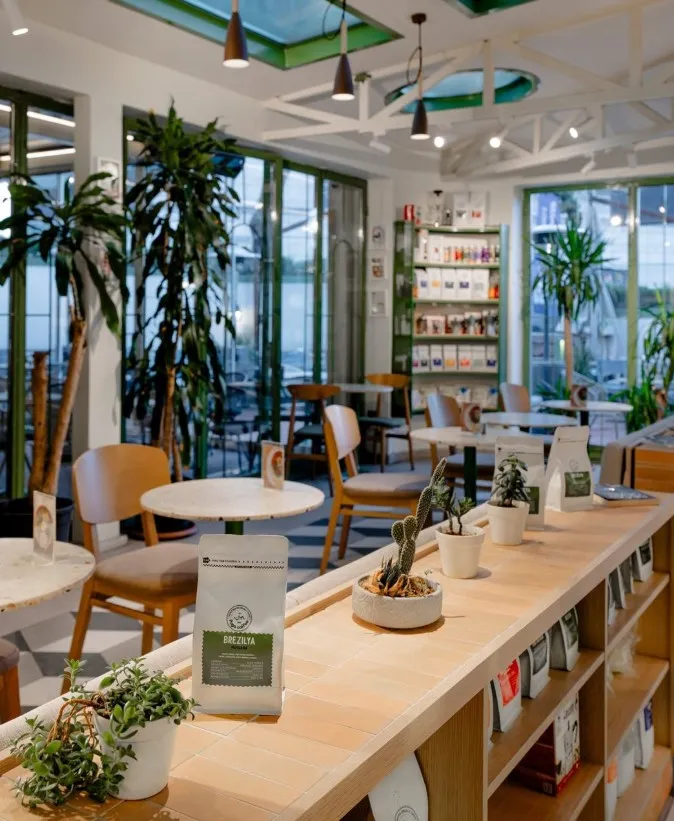 Yeşil çerçeveli geniş cam pencerelerden doğal ışık alan, modern mobilyaları ve raflarında Brezilya kahveleri bulunan ferah bir kafe iç mekanı, İstanbul'da verimli çalışmak için ideal çalışma mekânlarından biri olan Fuga Coffee'nin huzurlu ve estetik atmosferini yansıtıyor.