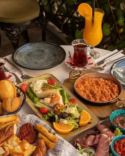 Dış mekanda, beyaz masa örtüsü serilmiş bir masada, ortasında çeşitli peynirler, domates, salatalık ve cevizden oluşan bir tabak, bakır tavada menemen, taze ekmekler, sucuk, patates kızartması ve çeşitli soslar içeren küçük mavi kaselerin yanı sıra, bir bardak portakal suyu ve ince belli bir çay bardağının yer aldığı zengin bir kahvaltı sofrası sunumu.