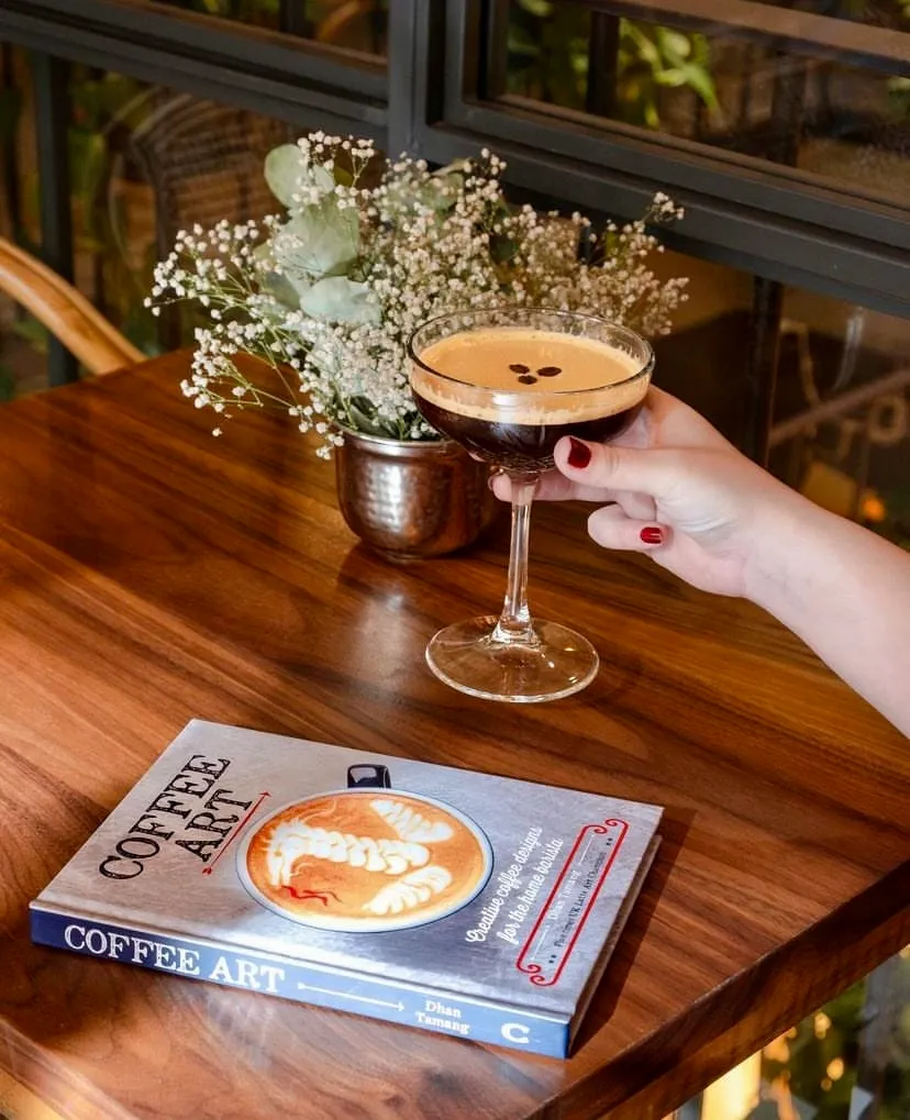 Koyu kırmızı ojeli bir elin tuttuğu, kremalı tepesinde üç kahve çekirdeği bulunan koyu renkli kadeh bir kahve kokteyli, sıcak tonlu ahşap bir masanın üzerinde Coffee Art kitabının ve küçük bir vazodaki beyaz çiçekler ile yeşil yaprakların yanında dururken, arka planda pencereden bulanık yeşil bir dış manzara görünüyor.