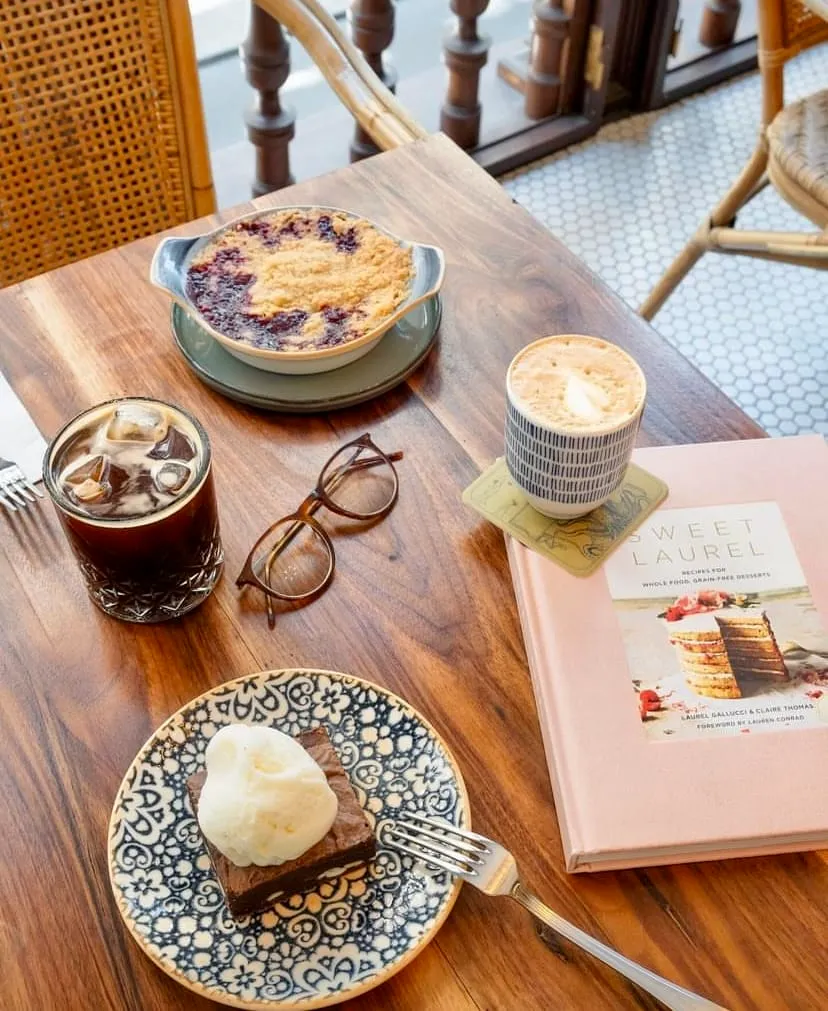 Ahşap bir masanın üzerinde, dondurmalı bir brownie, buzlu kahve, köpüklü bir latte, meyveli crumble, kahverengi çerçeveli gözlükler ve açık pembe bir yemek kitabı düzenli bir şekilde yer alırken, arka planda hasır sandalyeler ve ahşap korkuluklar görünen bir kafe köşesi.