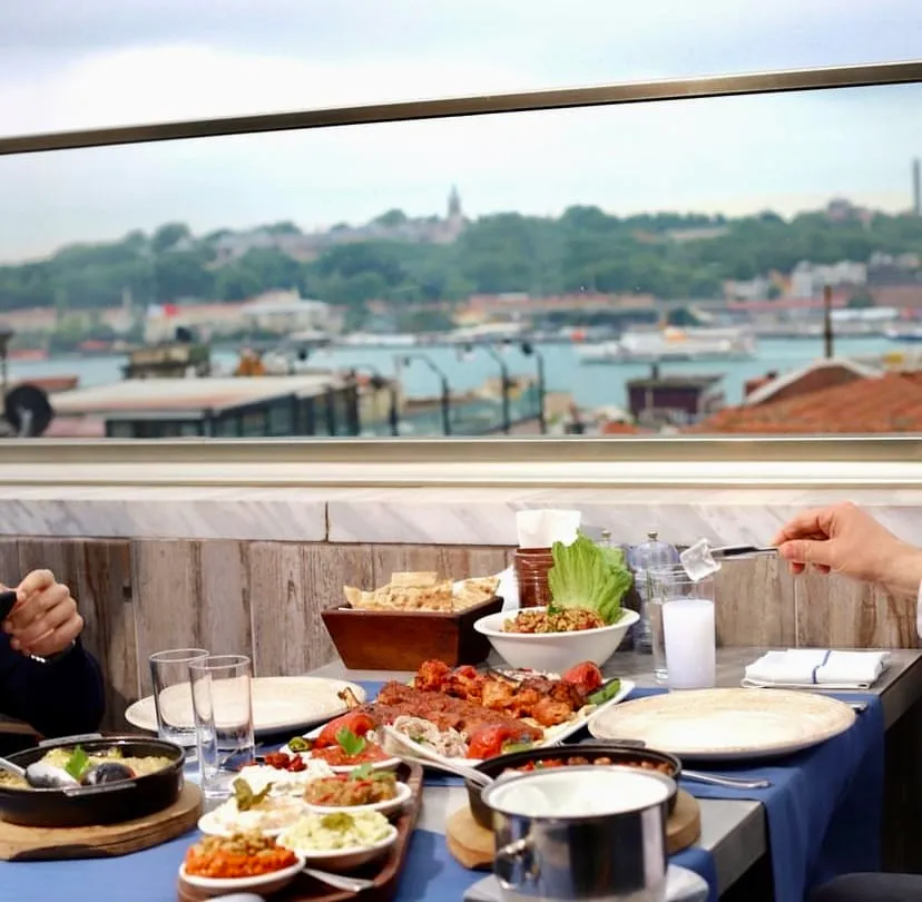 İstanbul'un tarihi yarımadasına nazır, cam korkuluklu bir terasta yer alan masada, çeşitli Türk mezeleri, ızgara etler ve ekmeklerle birlikte servis edilen rakıya buz ekleyen bir el görülüyor; arka planda Boğaz'ın ve şehrin etkileyici panoramik manzarası uzanıyor.
