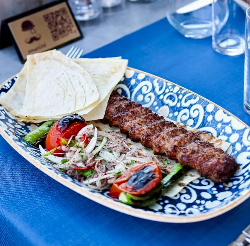 Mavi ve beyaz desenli bir servis tabağında, ızgarada pişirilmiş uzun bir Adana kebap, yanında ince lavaş ekmekleri, közlenmiş domates ve biberler ile sumaklı soğan salatası sergileniyor, arka planda bir restoran masası ve QR kodlu menü standı görülüyor.