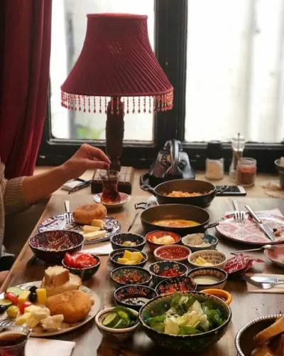 Kırmızı püsküllü bir abajurun aydınlattığı ahşap masada, çeşitli zeytinler, peynirler, reçeller ve soslarla dolu birçok küçük kaseden oluşan, çay eşliğinde sunulan zengin bir Türk kahvaltısı Payidar Galata'da göz alıcı bir şekilde sergileniyor.