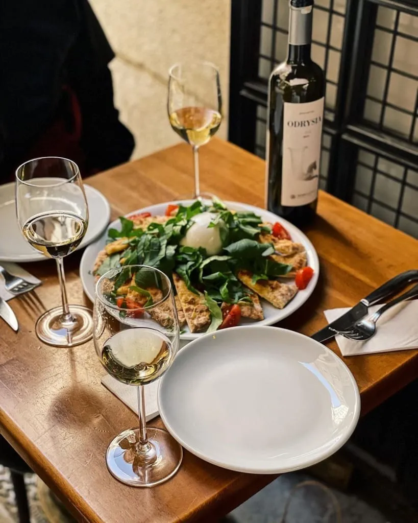Ahşap bir masada, üzerinde bol roka, çeri domates ve ortasında taze burrata peyniri bulunan, pizza veya pide tabanlı iştah açıcı bir yemek tabağı ile üç kadeh beyaz şarap ve bir şişe Odryses şarabı, keyifli bir pizzeria ambiyansını sergiliyor.