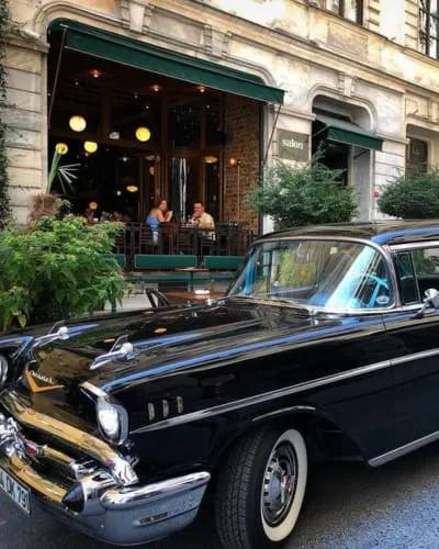 Ön planda parlak siyah, krom detaylı klasik bir otomobilin park ettiği, arka planda ise yeşil tenteli ve içinde müşterilerin oturduğu şık bir mekan olan Salon Galata'nın görüldüğü canlı bir şehir sahnesi.